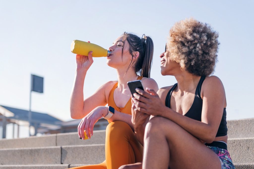 two female runner friends drinking water on a run break while using a mobile sitting on stairs outdoors, concept of friendship and sporty lifestyle, copy space for text